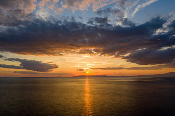 Vivid sunset with golden light reflecting across tranquil open water, dramatic clouds stretching overhead and distant silhouettes on the horizon.