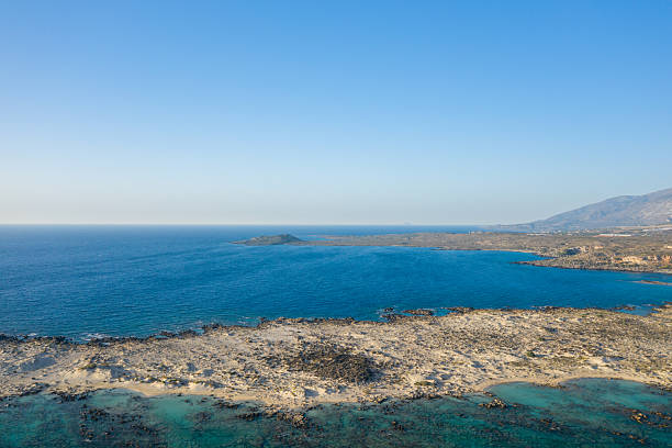 Expansive aerial view of a sandy, rocky shoreline meeting calm blue water at Elafonisi, Crete. Clear sky and gentle daylight create a serene and open coastal landscape.
