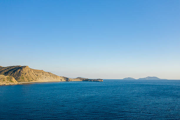 Expansive view of a tranquil blue bay with rugged coastal hills and distant islands under a clear sky on the southern coast of Crete.