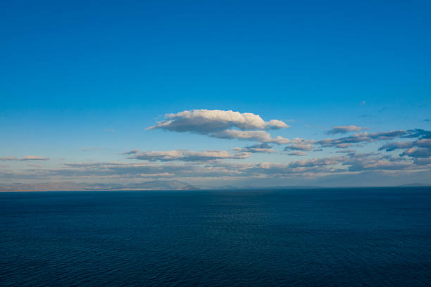 Vast expanse of deep blue water stretches toward a hazy mountain range on the horizon, beneath a sky scattered with fluffy clouds. The tranquil scene is bathed in soft daylight, evoking a sense of serenity and openness.
