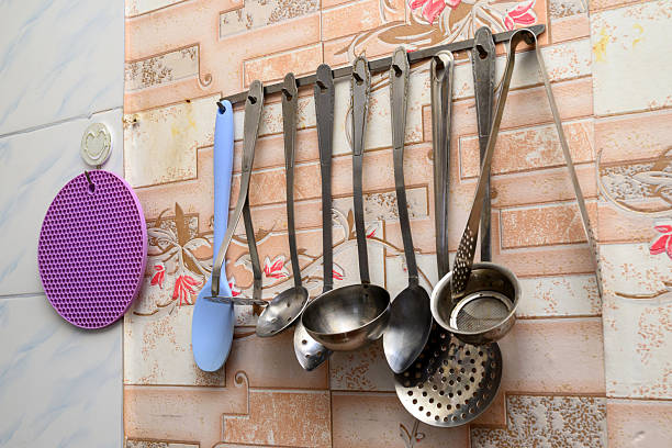 steel ladles and spatulas hang on a bar next to a round purple silicone hot pad fixed on a wall with beige tiles