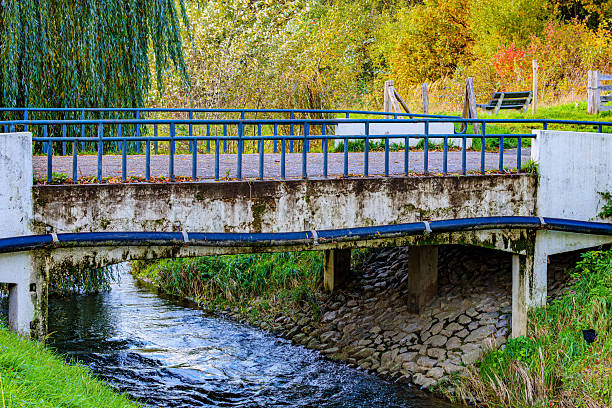 Small old vehicular bridge over Geleenbeek River, mossy walls, plastic pipe and metal fence, bench and yellowish-green trees in background, winter day in Munstergeleen, Limburg, Netherlands