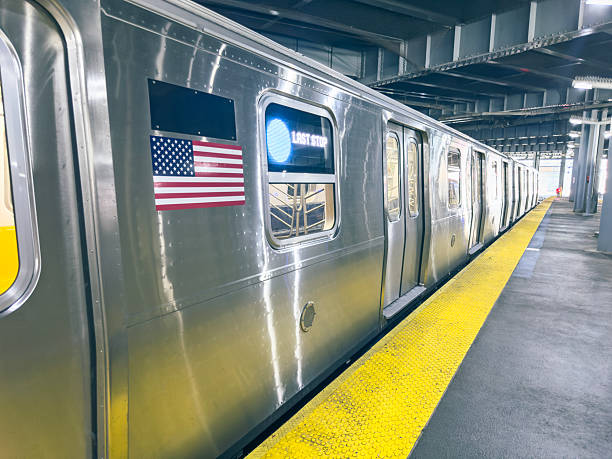 Side view of a stainless steel New York City subway train stopped at a clean, modern underground station platform with yellow safety edge, American flag on the car and overhead lighting, illustrating urban public transportation, daily commute and city infrastructure with no passengers in sight.​Ideal for use in articles and campaigns about NYC public transit, commuting, tourism, sustainable transportation, infrastructure investment, metro system guides, travel blogs and editorial stories about life in New York City.