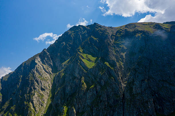 Steep rugged rock faces with patches of green grass rise dramatically under bright sunlight and scattered clouds in the Pyrenees mountains. Deep shadows accentuate the jagged textures and bold contours of the landscape.