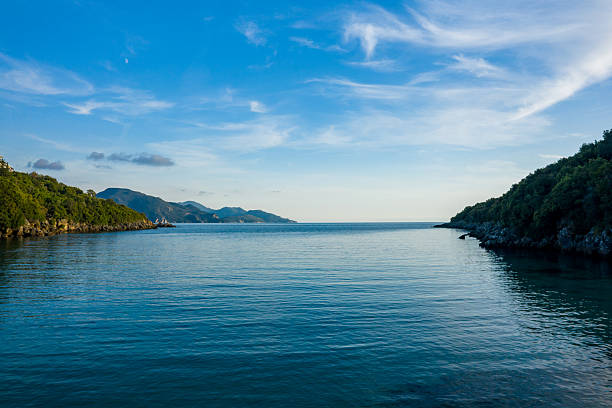 Tranquil bay with clear blue water framed by lush green hillsides at Syvota beach in Epirus, Greece. Soft evening light and wispy clouds create a peaceful coastal atmosphere with distant mountain silhouettes.