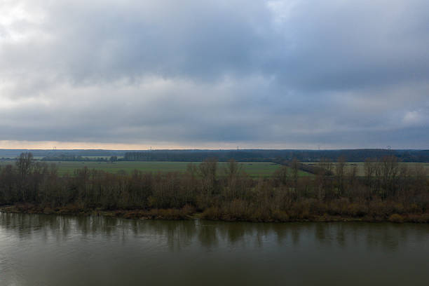 Clouds hang low over the Loire river, reflecting on calm water with leafless trees and green fields stretching into the distance. The subdued winter palette creates a peaceful, expansive mood.