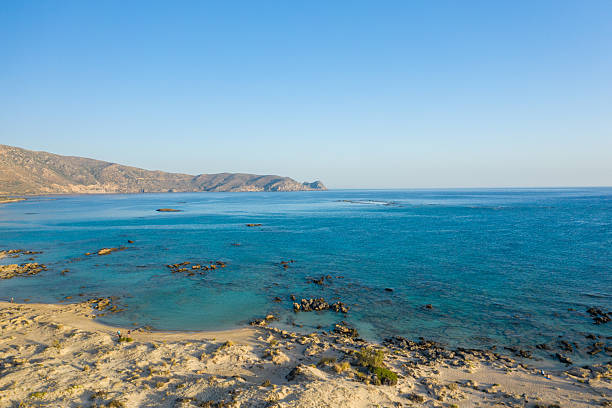 Wide view of turquoise shallow waters meeting a rocky shoreline at Elafonisi beach in Crete, Greece, with arid hills in the distance under a clear blue sky. The scene is sunlit, open, and naturally inviting.