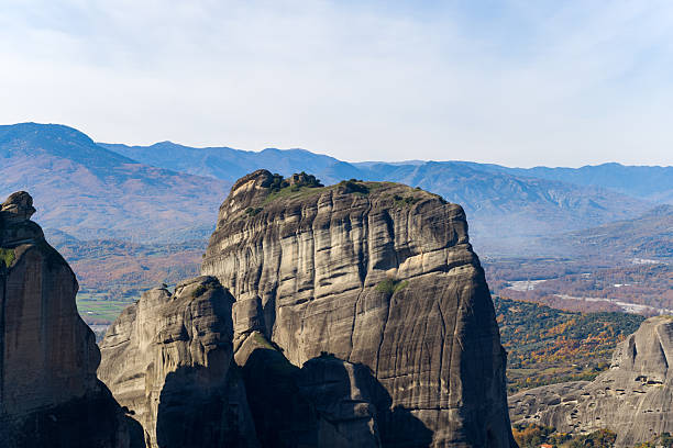A prominent sandstone monolith rises above the autumn landscape of Meteora, Greece, with layered textures illuminated by soft daylight and distant blue mountains in the background.