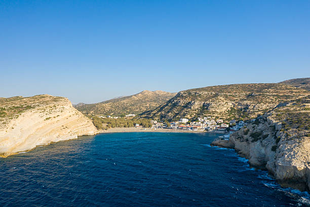 Vivid aerial view of Matala bay in Crete, showing turquoise water bordered by pale limestone cliffs and a small coastal village nestled among rugged hills under a clear blue sky.