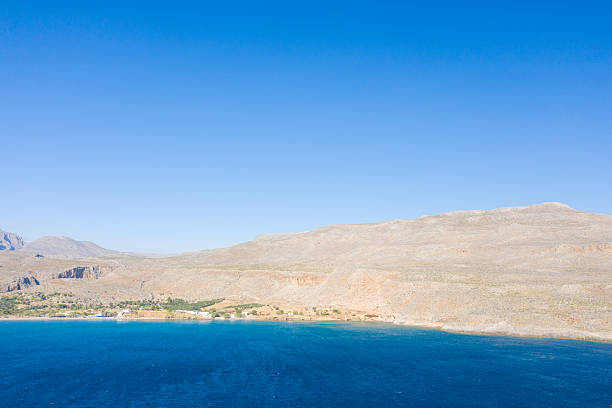 Bright blue water meets sunlit, rocky hills under a vast clear sky at Kato Zakros, Crete. The minimal shoreline and open landscape evoke a sense of calm and spaciousness.