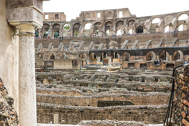 Detailed low-angle view of the Colosseum’s ancient arches and stone facade in Rome, Italy. The warm tones of the historic amphitheater contrast with the cloudy sky, emphasizing its monumental scale and timeless beauty.
