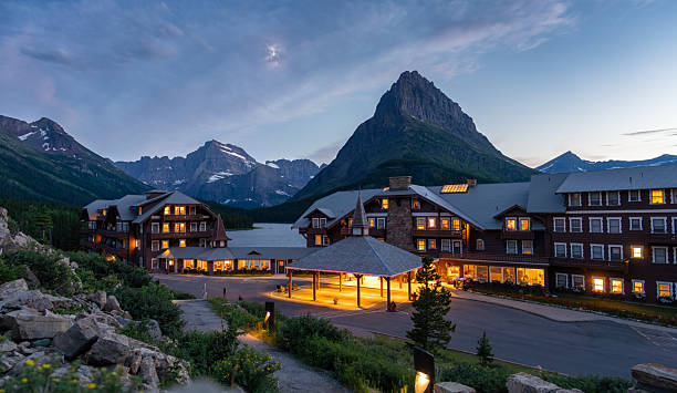 Many Glacier Hotel illuminated at dusk with the moon visible above dramatic alpine peaks in Glacier National Park, Montana, USA. Iconic mountain lodge during blue hour featuring warm lights, rugged Rocky Mountain scenery, and tranquil wilderness atmosphere, ideal for travel, tourism, hospitality, vacation, and destination concepts.