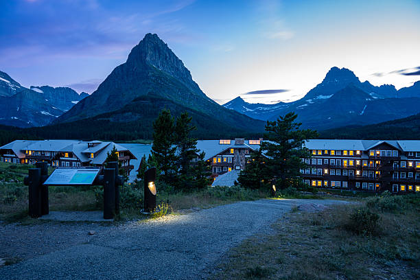Many Glacier Hotel illuminated at dusk below dramatic alpine peaks in Glacier National Park, Montana, USA. Iconic mountain lodge during blue hour featuring warm lights, rugged Rocky Mountain scenery, and tranquil wilderness atmosphere, ideal for travel, tourism, hospitality, vacation, and destination concepts.