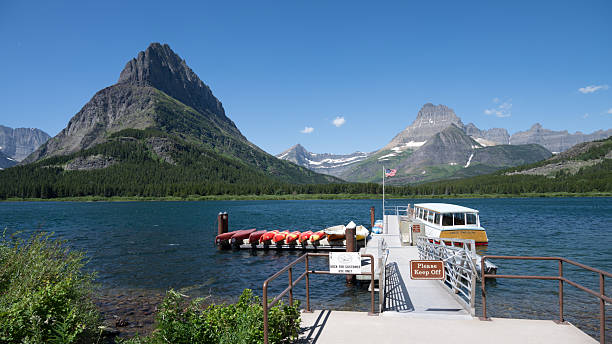Boat moored on a clear alpine lake beneath dramatic mountain peaks in Glacier National Park. Scenic summer landscape with rocky shoreline, turquoise water, evergreen forest, and rugged Rocky Mountain scenery, ideal for travel, tourism, wilderness, and nature concepts.
