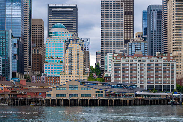 View of the Seattle waterfront with historic pier buildings in the foreground and the downtown high-rise skyline behind, photographed along Elliott Bay in Seattle. Modern office towers and residential buildings line the shoreline under overcast skies, illustrating urban development and city life in the Pacific Northwest.