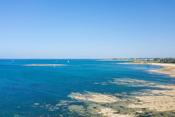 Expansive view of a clear tidal bay featuring rocky shallows, vibrant turquoise water, and a distant sandy shoreline under a cloudless blue sky near Anse de Trehennarvour in Saint Philibert.