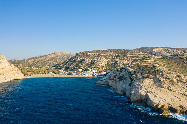 Aerial view of Matala on Crete, showing a small seaside village nestled between dramatic limestone cliffs and deep blue water, with sunlit hills in the background under a clear sky.