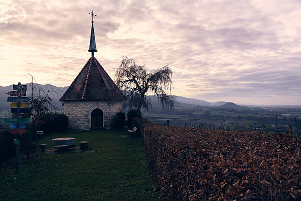 A small chapel stands on a hill surrounded by trees and bushes. The sky shows a mix of clouds during twilight. Mountains are visible in the background.