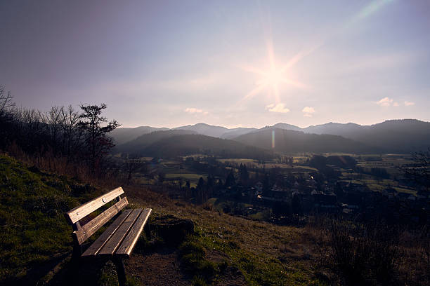 A wooden bench sits on a hilltop overlooking a valley at sunset. The sky is bright as the sun sets behind distant mountains. Trees surround the area, creating a nature scene for reflection.