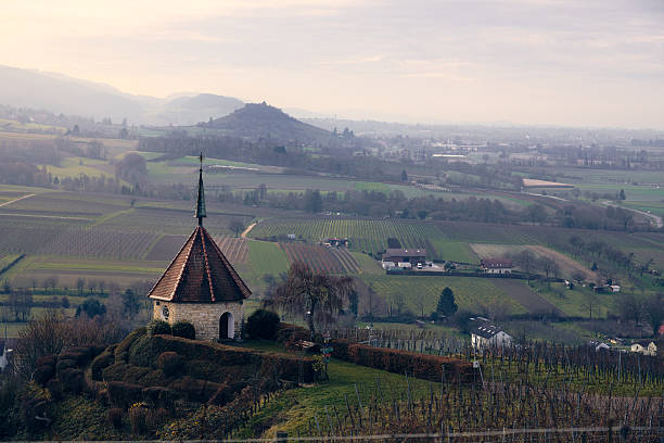 The sun rises over the hills, illuminating a small chapel on a hilltop. Green vineyards stretch out below, while distant landscapes fade into the horizon.