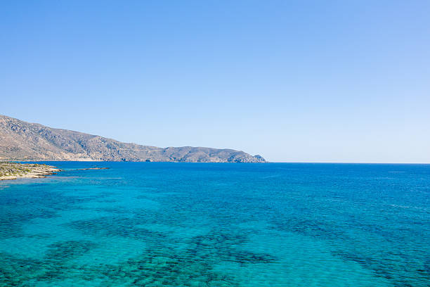 Expansive view of clear blue Mediterranean waters meeting a rocky shoreline and distant arid hills under a wide, cloudless sky. The tranquil seascape captures the vivid colors and open space of the Crete coastline.