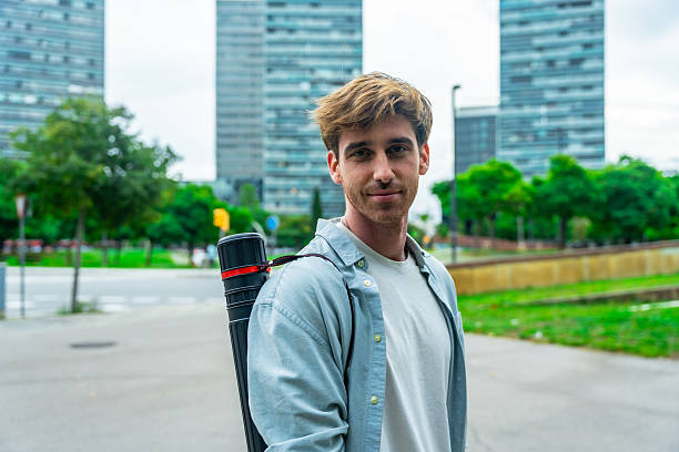 Young adult man smiling at camera, carrying a drawing tube on his shoulder, walking through a modern city park with residential skyscrapers in the background on an overcast day