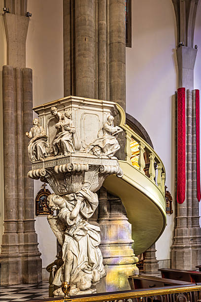 Catedral de San Cristobal de La Laguna, La Laguna, Spain, Europe - December 15th 2025:   An image of the exquisite Baroque pulpit carved in white Carrara marble, a prominent artwork inside the Cathedral of San Cristóbal de La Laguna on Tenerife. Created by the Italian sculptor Pasquale Bocciardo and completed in Genoa in 1767, the pulpit rests on a dramatic base featuring a kneeling angel or putto. The richly carved basket includes panels depicting the four Evangelists — Matthew, Mark, Luke and John — shown in dynamic, theatrical poses. An elegant curved staircase with a light green and gilded handrail ascends to the pulpit from the right. The artwork stands in striking contrast to the massive grey columns of the Neo‑Gothic nave. The church is rather new; the construction ended in 1915, and it is considered one of the first buildings in Spain to use concrete. It replaced an older, unsafe church. The new church inherited its status as a cathedral from the former building.