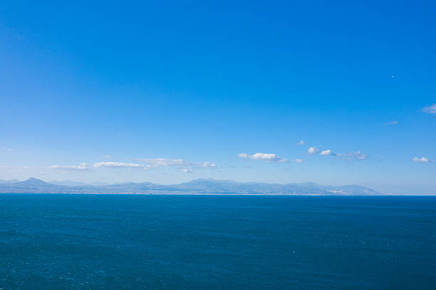 Wide open seascape featuring deep blue water and a faint mountain range on the horizon, all beneath a cloud-dotted, vibrant sky. The scene conveys spaciousness and calm, with crisp daylight enhancing the serene atmosphere.