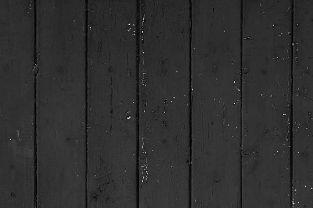 A close-up view of aged black wooden planks arranged vertically with visible grain, cracks, and chipped paint. The monochrome color palette and natural imperfections give the image a rustic, weathered appearance, suitable for use as a textured background or design element.