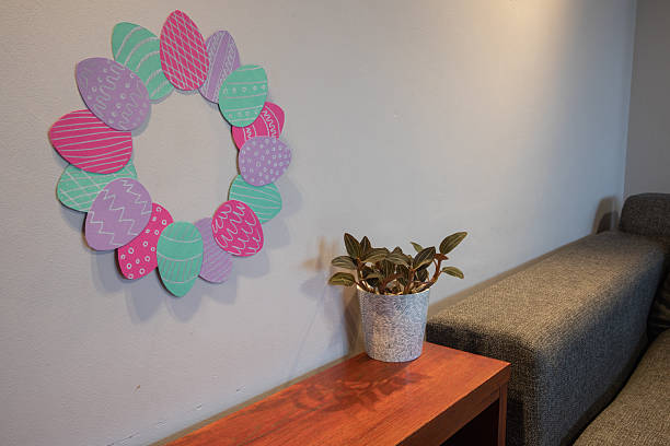 Handmade paper Easter egg wreath on a wall in a cozy modern interior with a houseplant and a grey sofa. Minimalist DIY festive home decoration.