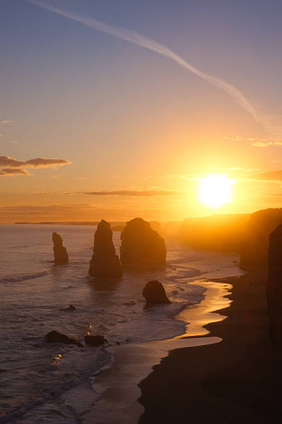 A breathtaking and serene vertical photograph capturing the magical sunset at the world-renowned Twelve Apostles, located along the scenic Great Ocean Road in Port Campbell National Park, Victoria, Australia. The sun, a brilliant orb of light, descends towards the horizon, bathing the sky in a spectacular gradient of deep blue, purple, and fiery orange. The iconic limestone sea stacks are cast in dramatic silhouette against the vibrant sky and the shimmering Southern Ocean. The golden light beautifully reflects on the wet sand of the beach, creating a luminous path that draws the viewer's eye into the peaceful scene.This high-quality, evocative image is perfect for projects related to travel, tourism, nature, landscapes, Australian destinations, and coastal beauty. It conveys concepts of tranquility, majesty, exploration, the end of a day, and natural wonders. Its strong vertical orientation makes it ideal for magazine covers, book covers, posters, website hero images, travel blogs, and compelling social media stories.