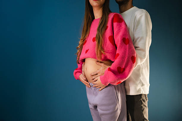 Studio photo of a young couple standing together with hands on a small baby bump. Represents early pregnancy, anticipation, and family beginnings.