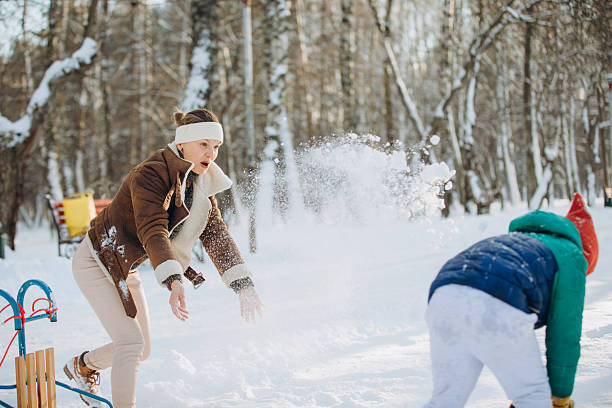Cheerful young couple laughing and throwing snowballs in a sunlit winter park, playful outdoor recreation in warm coats and mittens amid snowy trees and bright cold air