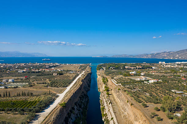 Wide aerial view of the Corinth Canal stretching toward the vivid blue sea, bordered by rural farmland, olive groves, and distant mountains beneath a bright, clear sky.