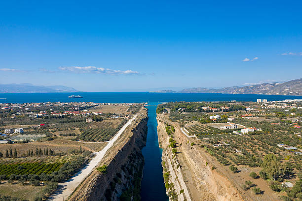 Aerial view of the Corinth Canal cutting through rural farmland and olive groves, leading to the deep blue sea with distant mountains under a clear sky.