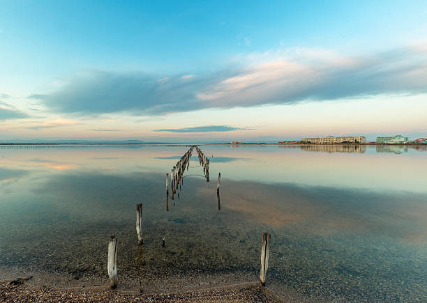 Аrranged weathered wooden poles in Pomirie's salt lake, buildings at the distance and reflection of the sky in the water. Bulgaria, Eastern Europe.