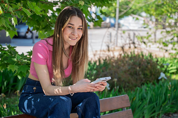 istock Young woman using phone sitting on park bench 2255367888