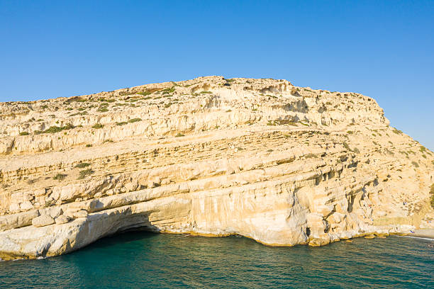 Bright sunlight highlights the layered limestone cliffs above a deep blue sea and a natural cave along the Matala coastline in Crete. The rugged rock formations and vivid sky create a striking Mediterranean landscape.
