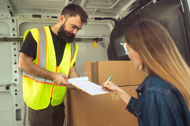 A female customer signs a clipboard held by a delivery courier standing inside a delivery van. The courier wears a high-visibility safety vest, documenting the completed handover.