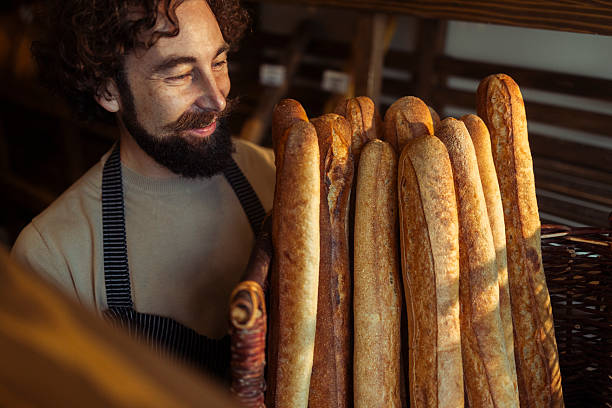 Cheerful male baker wearing black gloves carries a wicker basket overflowing with freshly baked baguettes in a traditional bakery, surrounded by shelves filled with various breads