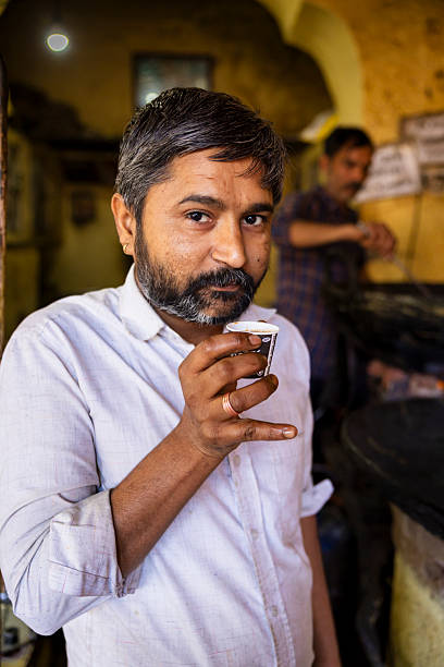 A middle-aged man with salt-and-pepper hair and a short beard holds a tiny paper cup of hot tea close to his lips, meeting the camera with a calm, knowing expression. He wears a light-colored, rolled-sleeve shirt and a simple ring; the warm, dim interior behind him shows another worker blurred in the background and large cooking vessels, giving the scene a candid, intimate tea-stall atmosphere.