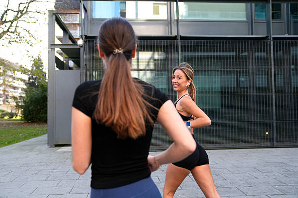 Two women running outdoors in a city park. One woman looks back at her friend smiling, both wearing activewear