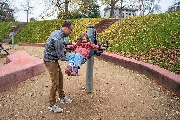 Man supporting his child on a rotating metal structure at a playground. Outdoor exercise and together time.