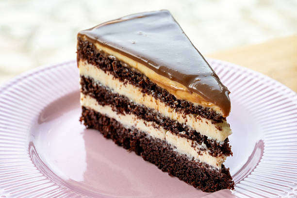 A decadent slice of chocolate layer cake with creamy white filling and a glossy caramel glaze, served on a ridged pink plate.
