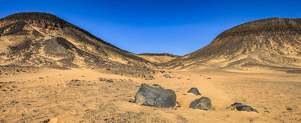 A wide, sunlit desert panorama showing golden sand plains strewn with dark basalt boulders leading toward rounded, black-capped hills. Wind-carved ripples and scattered stones mark the dunes, while stark contrast between the warm sand and rough volcanic rock creates a dramatic, otherworldly landscape beneath a clear deep-blue sky.