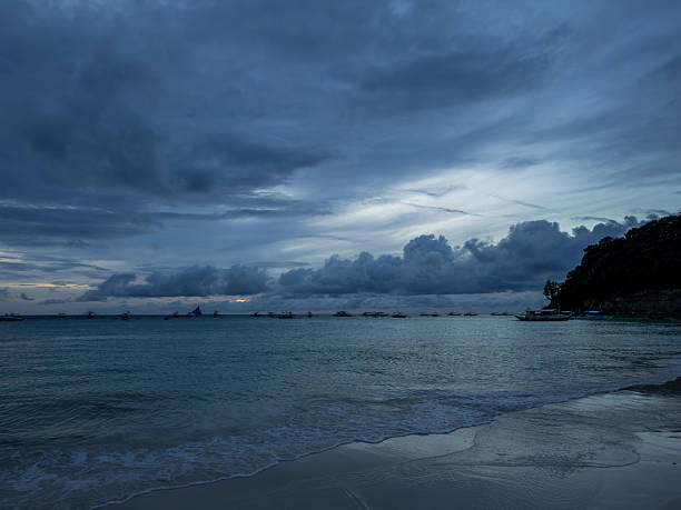 Moody twilight scene over a tropical beach in Boracay, Philippines. Dark clouds loom above tranquil waters and a sandy shore, with traditional boats silhouetted against the horizon. Serene and