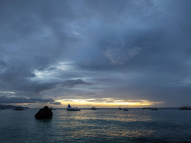 Dramatic sunset over Boracay's White Beach, featuring iconic Willy's Rock and traditional Filipino outrigger boats (banca) silhouetted against a cloudy, golden horizon.