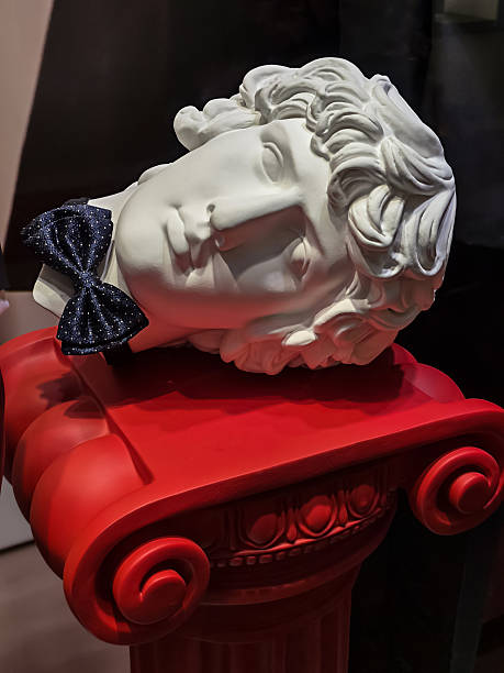 Classic plaster bust of young man wearing modern bow tie rests on bright red pedestal. Gothic elegance: fashion on edge of life and death.