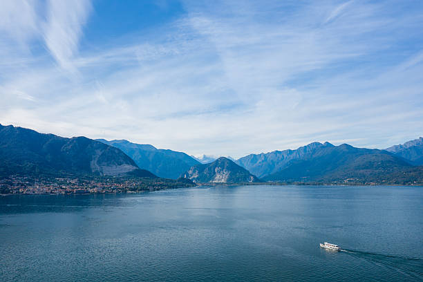 A white ferry moves across the tranquil blue waters of Lake Maggiore in northern Italy, set against a dramatic backdrop of alpine peaks and a sky streaked with wispy clouds. The expansive scene highlights the vast lake, distant shoreline, and rugged mountain landscape.