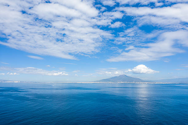 Expansive view of Mount Vesuvius rising above the distant shoreline, with calm blue waters of the Bay of Naples in the foreground and dramatic cloud formations filling the sky. The scene captures the iconic Italian coastline under bright daylight.